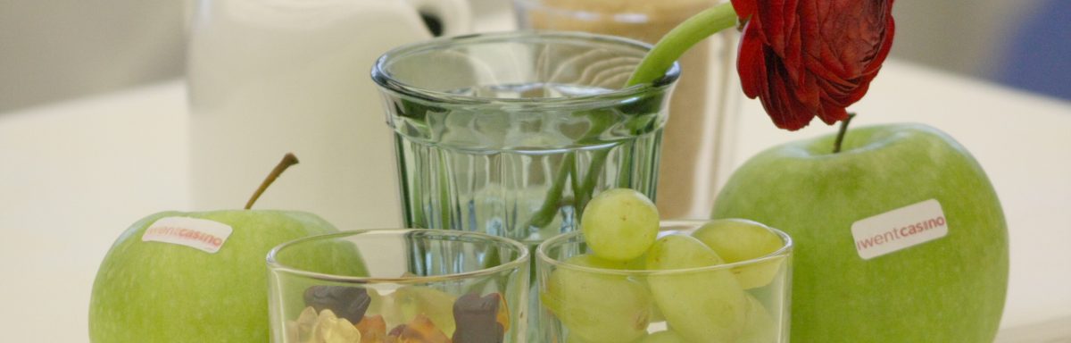 Zwei grüne Äpfel mit iwentcasino-Aufschrift und ein Glas Gummibärchen neben einem Glas Weintrauben, dahinter eine Blume im Glas